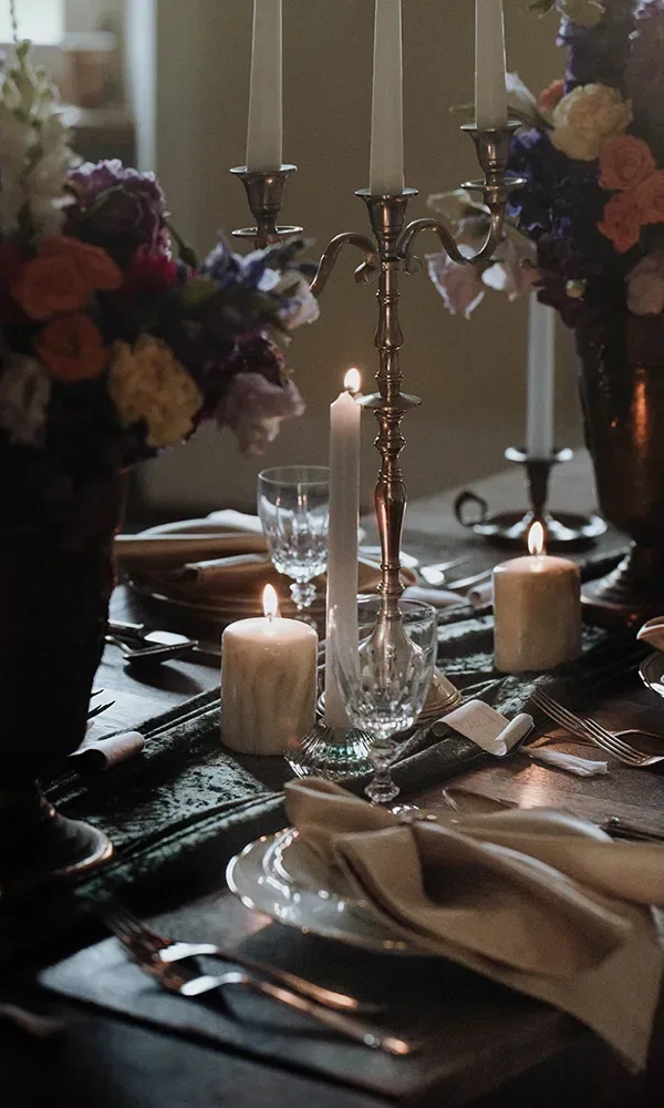 Ein festlich gedeckter Tisch mit brennenden Kerzen und hohen silbernen Kerzenständern, umgeben von Blumenarrangements. Gläser, Besteck und Servietten sind auf dem Holztisch kunstvoll angeordnet.