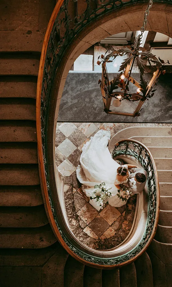 Luftaufnahme eines Hochzeitspaares auf einer Wendeltreppe, das Paar steht auf einem Mosaikboden, während die Braut einen voluminösen weißen Schleier trägt. Ein Kronleuchter hängt über ihnen.