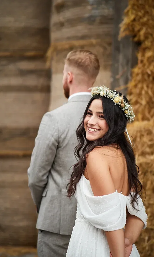Eine lächelnde Braut mit Blumenkranz im Haar schaut über die Schulter, während ihr Bräutigam mit dem Rücken zur Kamera steht. Die beiden befinden sich vor einem rustikalen Hintergrund aus Heuballen.