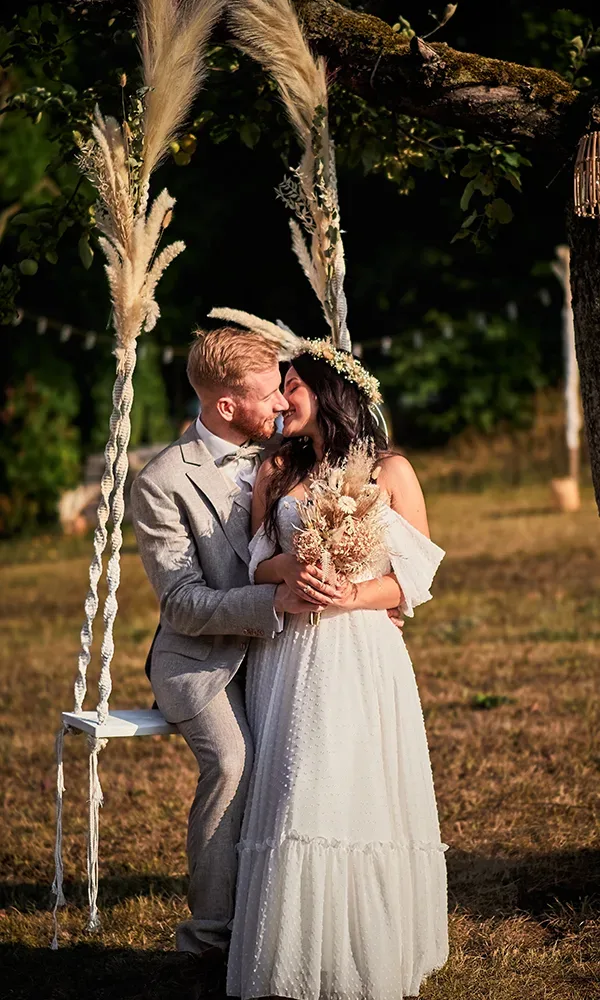 Ein Hochzeitspaar sitzt auf einer schwebenden Schaukel, geschmückt mit Pampasgras. Die Braut trägt einen Blumenkranz und hält einen Trockenblumenstrauß, während das Paar liebevoll miteinander interagiert.
