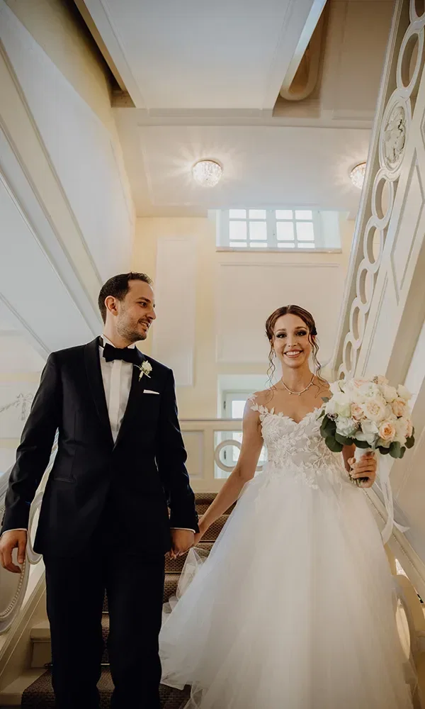 Das Brautpaar, der Bräutigam in einem schwarzen Anzug und die Braut in einem weißen Kleid, geht Hand in Hand eine elegante Treppe hinunter. Die Braut hält einen Brautstrauß mit weißen und rosa Blumen, während beide lächelnd in die Kamera schauen.