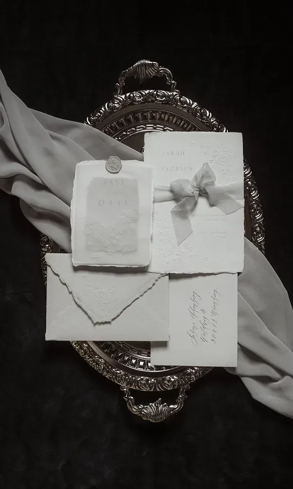Auf einem silbernen Tablett liegen elegante Hochzeitseinladungen und Save-the-Date-Karten, kunstvoll arrangiert. Eine graue Stoffserviette ist dekorativ um das Tablett drapiert.