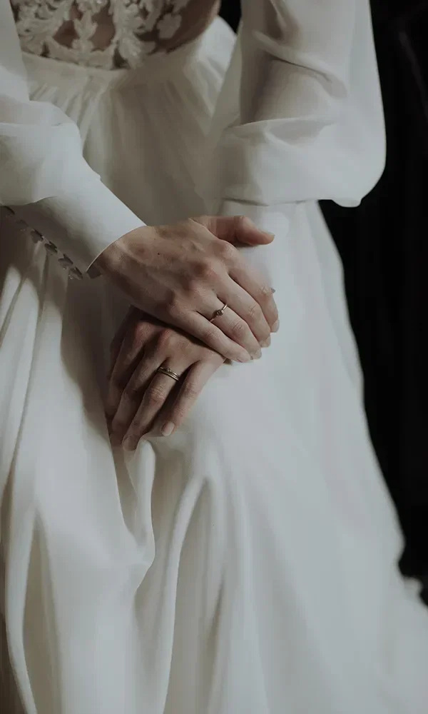 Nahaufnahme der Hände einer Braut, die übereinandergelegt sind. Sie trägt einen schlichten goldenen Ehering, und das weiße, fließende Brautkleid ist im Hintergrund sichtbar.
