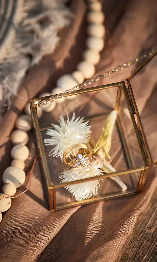 Zwei goldene Eheringe liegen in einer kleinen Glasbox, dekoriert mit weißen Trockenblumen. Die Box steht auf einem weichen, beigen Stoff, umgeben von einer Holzkette und weiteren Dekorationselementen.
