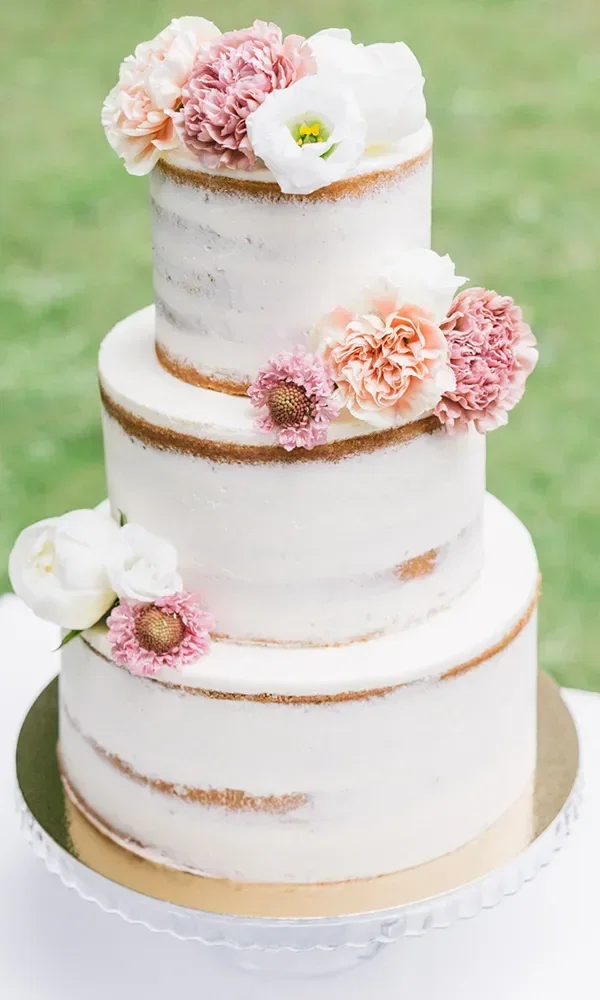 Eine dreistöckige Semi-Naked Hochzeitstorte mit dünner Buttercreme, verziert mit rosa und weißen Blumen, steht auf einem silbernen Tortenständer im Freien.
