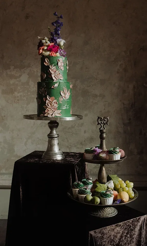 Eine dreistöckige grüne Hochzeitstorte mit floralen Verzierungen steht auf einem silbernen Tortenständer. Daneben sind auf einer Etagere Cupcakes und Früchte wie Trauben und Äpfel dekorativ arrangiert.