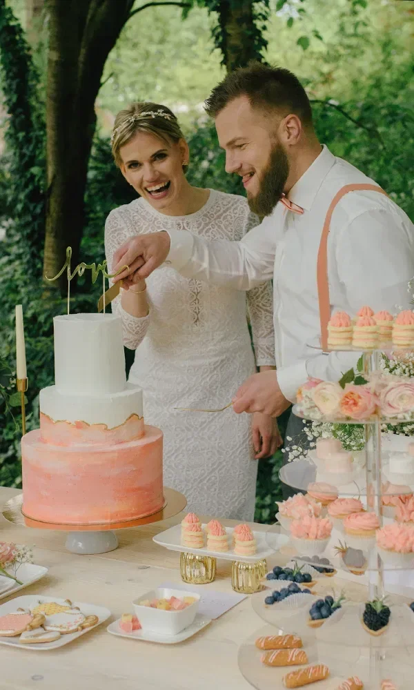 Ein Brautpaar schneidet gemeinsam eine zweistöckige Hochzeitstorte an. Die Torte ist weiß und rosa gefärbt, während das Paar sichtlich Freude am Moment hat. Im Hintergrund steht ein Tisch mit weiteren Süßspeisen.