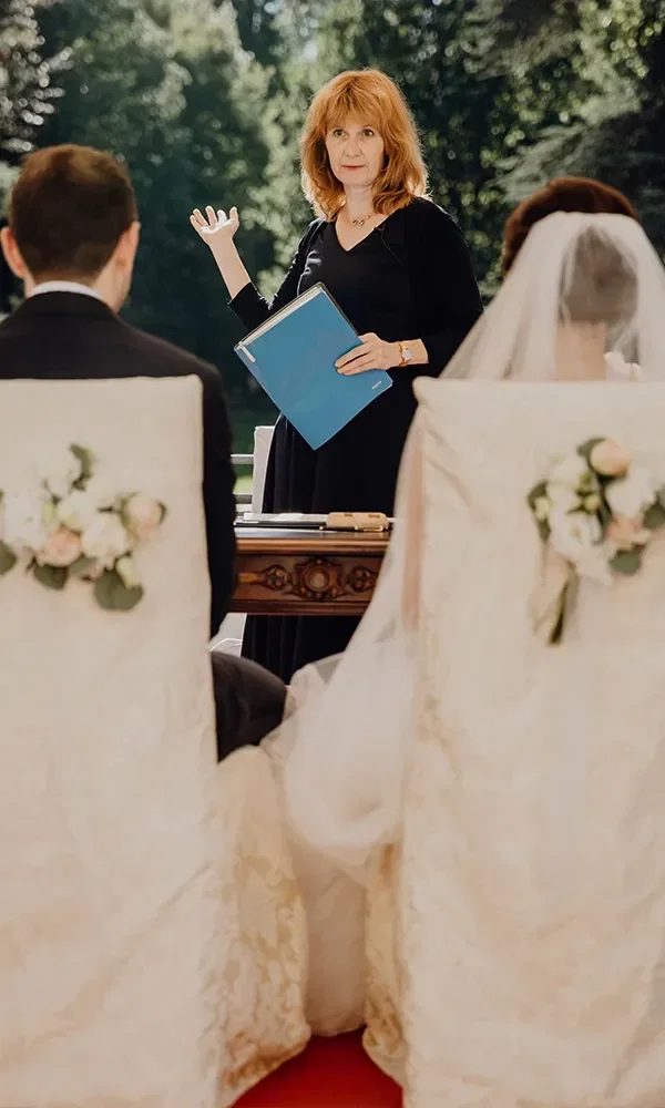 Eine Standesbeamtin hält eine Hochzeitszeremonie im Freien ab, während das Brautpaar mit dem Rücken zur Kamera sitzt. Beide Stühle sind mit Blumen dekoriert, und die Beamtin hält ein blaues Buch in der Hand.
