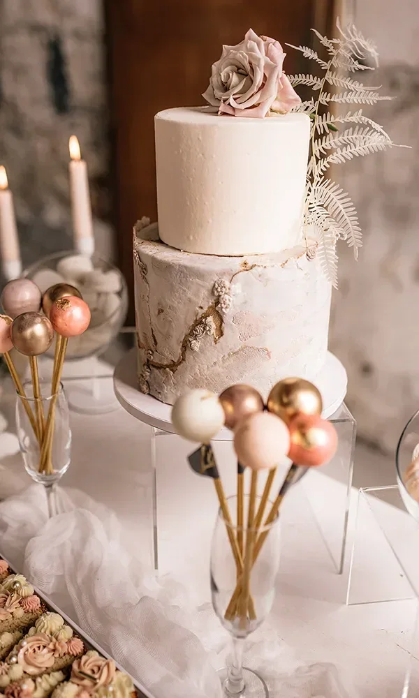 Eine zweistöckige Hochzeitstorte, dekoriert mit einer großen Blume und filigranen Blättern, steht auf einem Tisch. Neben der Torte befinden sich bunte Cake-Pops in Gläsern und weitere süße Dekorationen auf einem weißen Tischtuch.