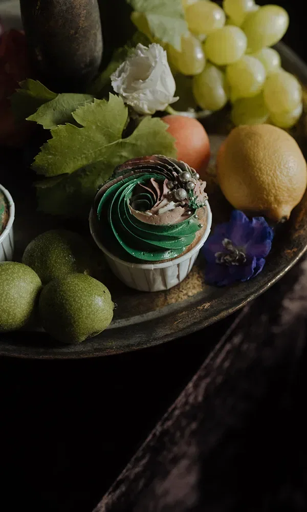 Ein Teller mit essbarer Dekoration, darunter ein Cupcake mit grünem und braunem Zuckerguss, umgeben von Trauben, Limetten und einer Zitrone. Blumen und Blätter verzieren die Anordnung.