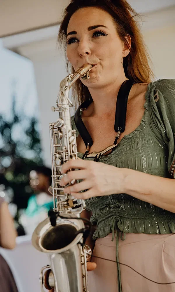 Eine Frau in einem grünen Oberteil spielt Saxophon während einer Hochzeitsfeier. Sie konzentriert sich auf ihre Musik, während sie das Instrument mit beiden Händen hält.