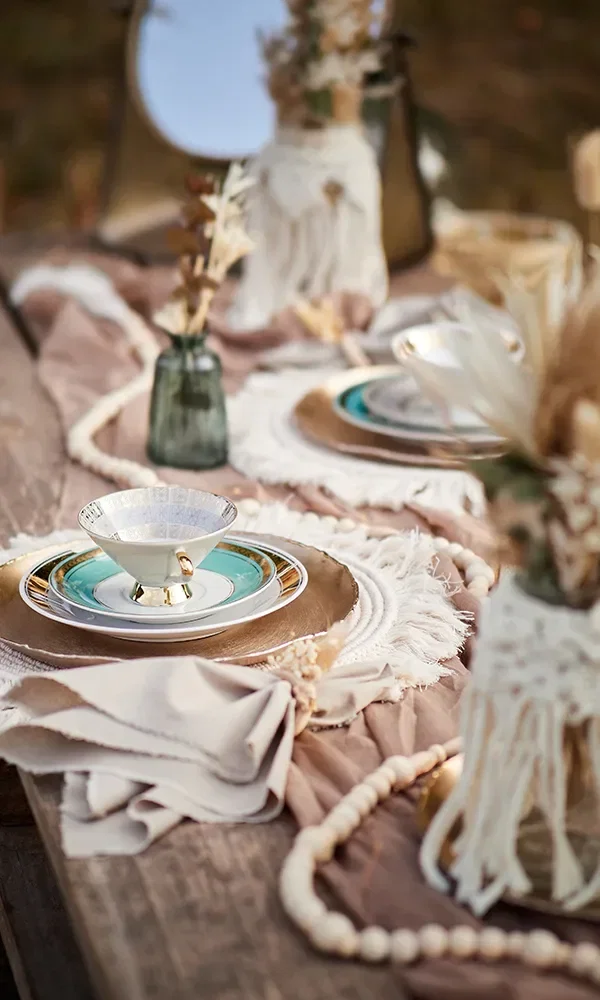 Ein festlich gedeckter Tisch im Freien mit einer goldenen Servierplatte, darauf liegt elegantes Porzellan mit einer grün-goldenen Teetasse. Der Tisch ist mit naturfarbenen Stoffen, Trockenblumen in kleinen Vasen und Holzperlen dekoriert.