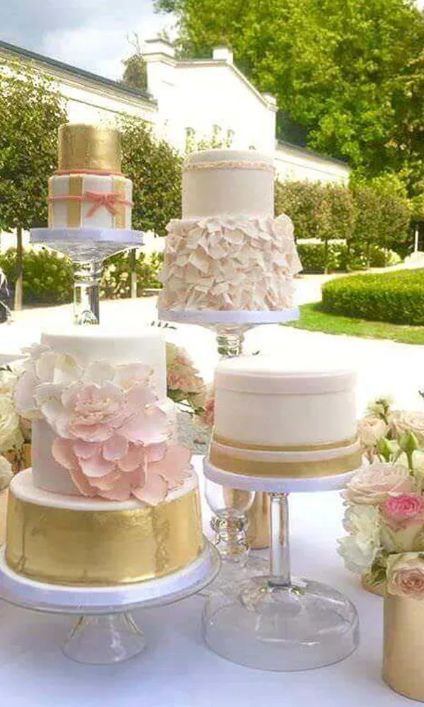 Eine Auswahl verschiedener Hochzeitstorten, jede mit unterschiedlichen Designs und goldenen Details, steht auf einem Tisch im Freien. Die Torten sind mit Blumen und eleganten Verzierungen dekoriert.