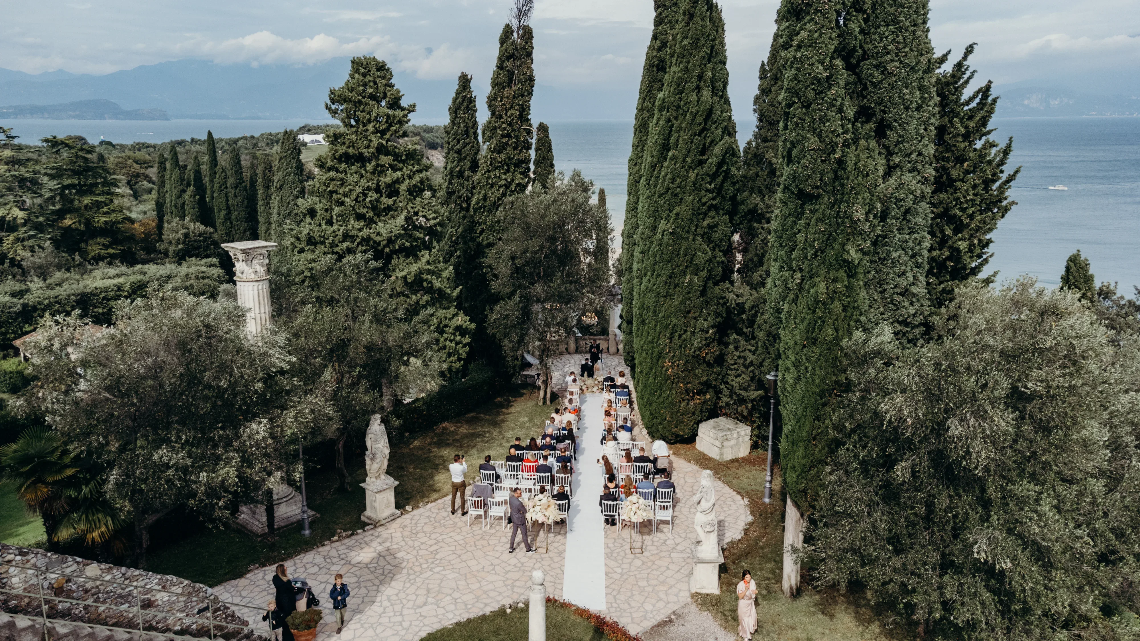 Eine Hochzeit im Freien mit Blick auf das Meer, umgeben von hohen Zypressen und antiken Statuen, mit Gästen, die entlang eines gepflasterten Pfades sitzen.