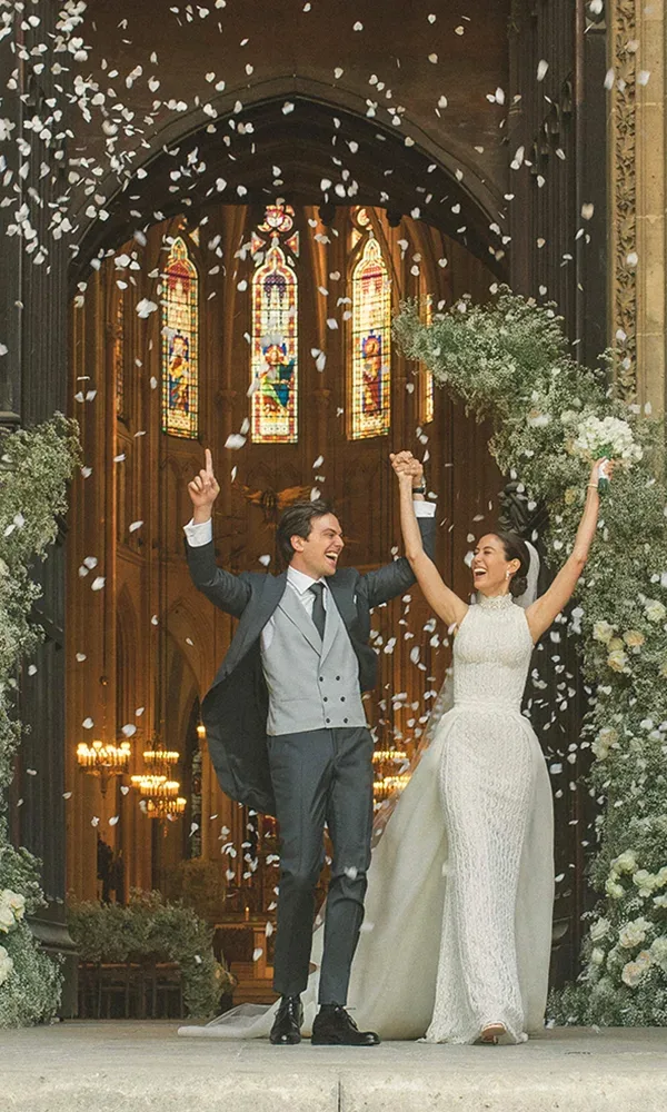 Das Brautpaar steht jubelnd vor einer großen Kirche, die in warmem Licht erstrahlt, während Konfettiblütenblätter um sie herumwirbeln. Die Braut trägt ein elegantes weißes Kleid, und der Bräutigam einen klassischen Anzug.