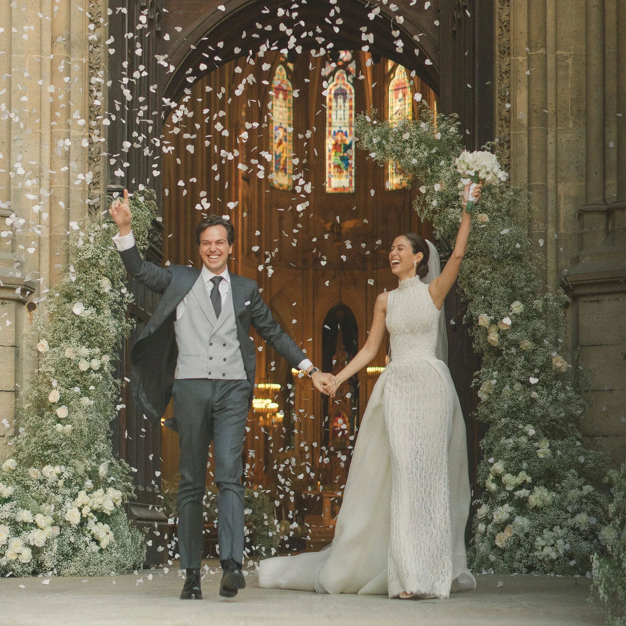 Ein frisch verheiratetes Brautpaar verlässt glücklich die Kirche, während Blütenblätter auf sie herabregnen. Beide halten die Hände in die Luft, strahlend vor Freude, umgeben von üppiger Blumendekoration.
