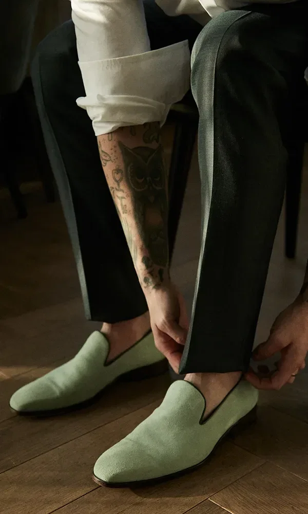Ein Mann mit tätowiertem Arm zieht sich grüne Slipper an, während er auf einem Holzboden sitzt. Seine Hosen sind dunkel, und der Lichtschein fällt sanft auf den Boden.