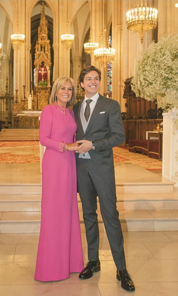 Der Bräutigam steht mit seiner Mutter, die ein elegantes, pinkfarbenes Kleid trägt, in einer prachtvoll geschmückten Kirche. Beide lächeln und halten sich an den Händen vor einem reich verzierten Altar.