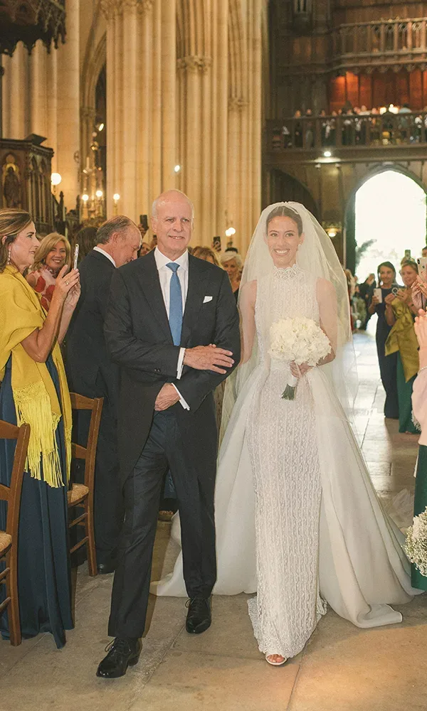 Eine Braut im eleganten weißen Kleid und mit einem Brautstrauß aus weißen Blumen wird von ihrem Vater in einer majestätischen Kirche zum Altar geführt. Die Gäste stehen auf und schauen dem Paar lächelnd zu.