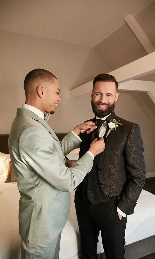 Der Trauzeuge richtet die Fliege des Bräutigams, der einen dreiteiligen dunklen Anzug trägt. Beide Männer wirken entspannt und lachen, während sie sich in einem modern eingerichteten Zimmer auf die Hochzeit vorbereiten.