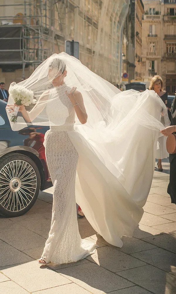 Eine Braut in einem eleganten, hochgeschlossenen weißen Brautkleid mit langem Schleier steigt aus einem Auto und hält einen Brautstrauß. Der Wind fängt ihren Schleier, der majestätisch hinter ihr weht.