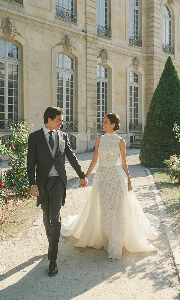 Ein Hochzeitspaar spaziert Hand in Hand vor einem eleganten Schloss entlang. Die Braut trägt ein hochgeschlossenes, weißes Kleid mit einem langen, fließenden Rock, während der Bräutigam in einem eleganten Anzug gekleidet ist.