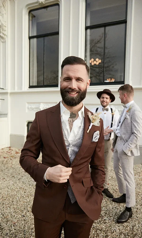 Der Bräutigam steht lächelnd in einem braunen Anzug und einer silbernen Weste vor einem weißen Gebäude. Im Hintergrund stehen zwei seiner Groomsmen, die in hellen Anzügen und Accessoires wie Hüten und Hosenträgern die entspannte Hochzeitsatmosphäre genießen.