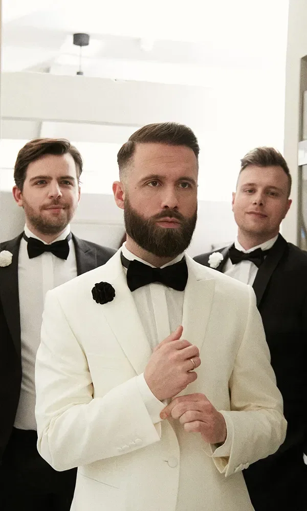 Der Bräutigam, in einem weißen Smoking mit schwarzer Fliege, steht selbstbewusst mit zwei seiner Groomsmen, die klassische schwarze Anzüge tragen. Alle wirken konzentriert und bereit für den großen Moment der Hochzeit.