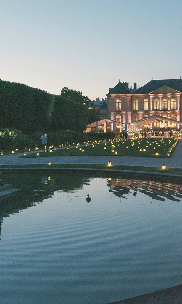 Ein beleuchtetes Schloss in der Abenddämmerung mit einem ruhig daliegenden Teich im Vordergrund. Der Garten ist mit zahlreichen Kerzen geschmückt, die den Weg zum Eingang des Schlosses sanft erhellen.