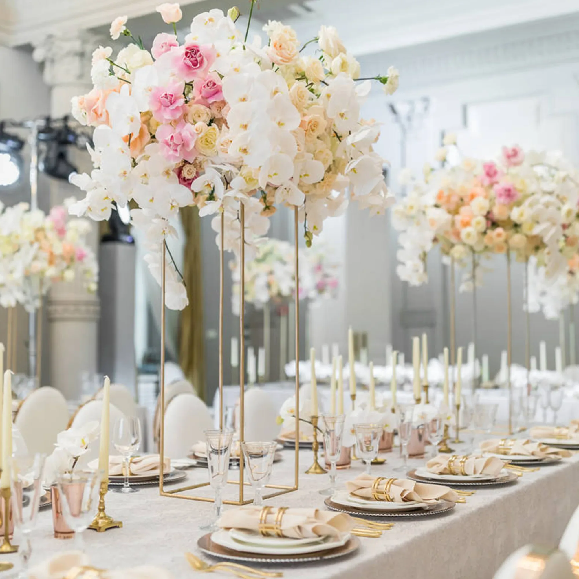 Eine festlich gedeckte Hochzeitstafel mit hohen Blumenarrangements in Pastellfarben. Die Tafel ist mit goldenen Kerzenhaltern, gläsernen Tellern und edlen Stoffservietten dekoriert, die in goldene Serviettenringe gewickelt sind.