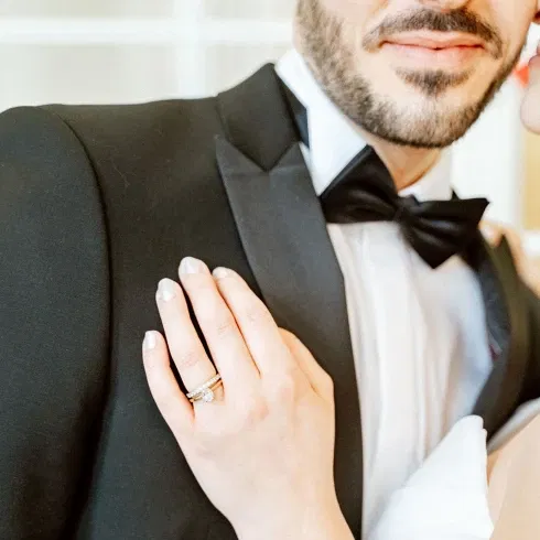 Nahaufnahme einer Hand der Braut, die sanft auf die Schulter des Bräutigams in einem schwarzen Smoking gelegt ist. Ihr eleganter Verlobungsring mit Diamanten ist im Fokus.