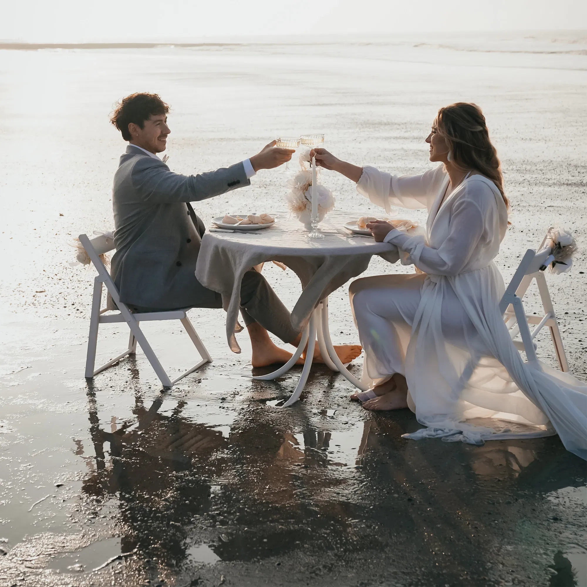 Hochzeitspaar an einem runden Tisch am Strand, das mit Champagner auf ihre Ehe anstößt. Beide sitzen barfuß und genießen den romantischen Moment im Abendlicht.
