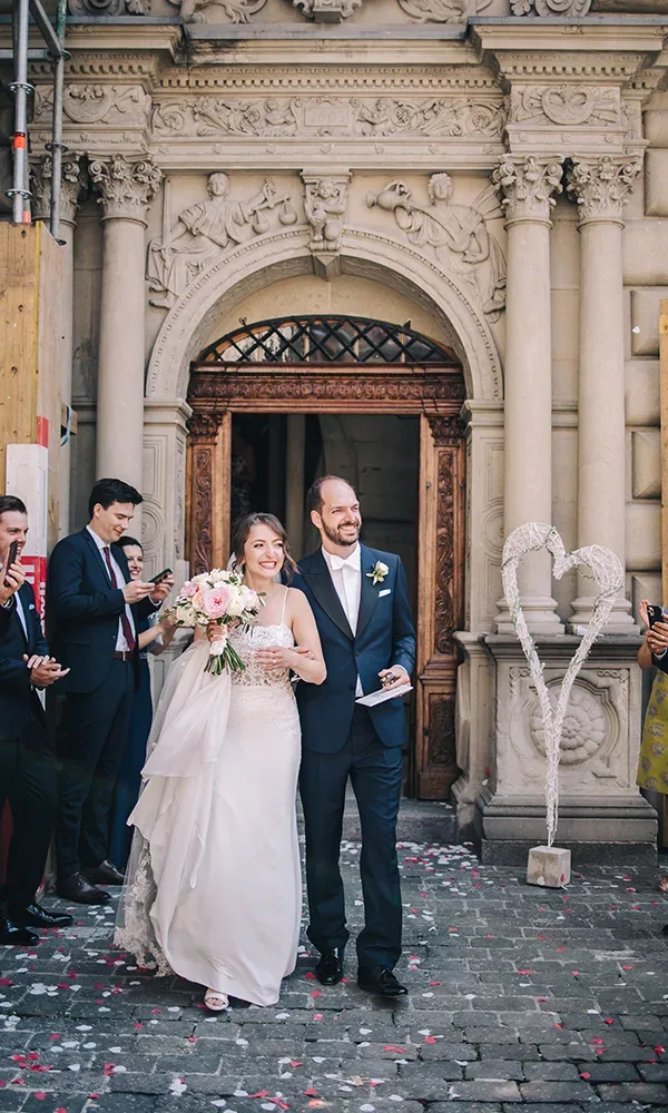 Ein frisch verheiratetes Paar verlässt strahlend ein prunkvolles Gebäude. Die Braut trägt ein weißes Kleid und hält einen Blumenstrauß, während der Bräutigam einen eleganten dunklen Anzug trägt. Die Gäste stehen jubelnd und applaudierend daneben.