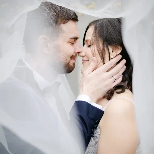 Ein Brautpaar steht eng beieinander, lächelnd und zärtlich, während der Schleier der Braut sie umhüllt und ein intimer Moment zwischen den beiden entsteht.