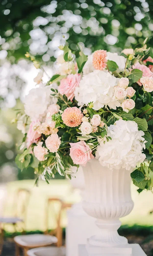 Ein großer, üppiger Blumenstrauß in einer weißen Vase, bestehend aus weißen und rosafarbenen Blüten, darunter Hortensien und Rosen. Die Vase steht im Freien unter Bäumen, was eine romantische Atmosphäre schafft.