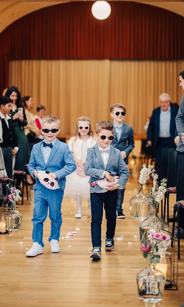 Mehrere Kinder in festlicher Kleidung, die während einer Hochzeitszeremonie den Gang entlang laufen. Die Jungen tragen Sonnenbrillen und Anzüge, während sie Stofftiere halten. Im Hintergrund folgen ein Blumenmädchen und weitere Gäste.
