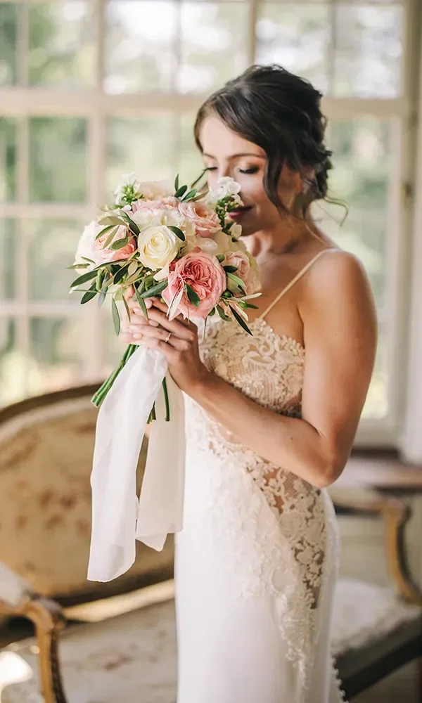 Eine Braut in einem zarten, ärmellosen Kleid hält einen duftenden Blumenstrauß aus rosafarbenen und weißen Blumen, während sie verträumt den Strauß betrachtet. Im Hintergrund ist ein hell erleuchteter Raum mit großen Fenstern zu sehen.