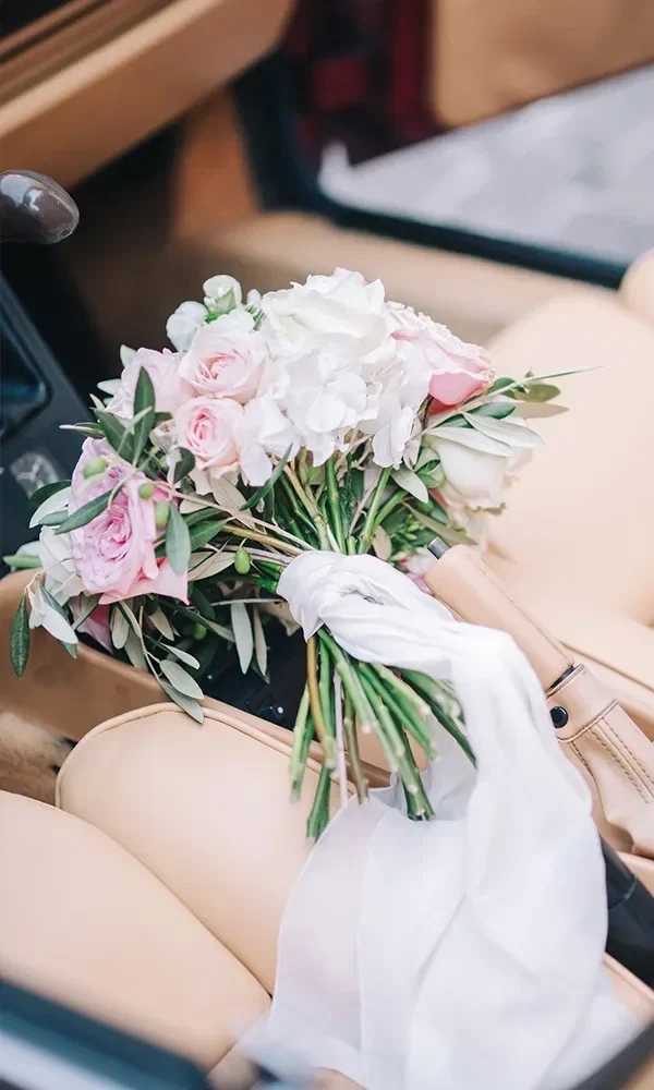 Ein schöner Brautstrauß aus weißen und rosafarbenen Blumen, arrangiert auf einem Autositz. Der Strauß ist mit einem weißen Band verziert und liegt auf beigen Ledersitzen.