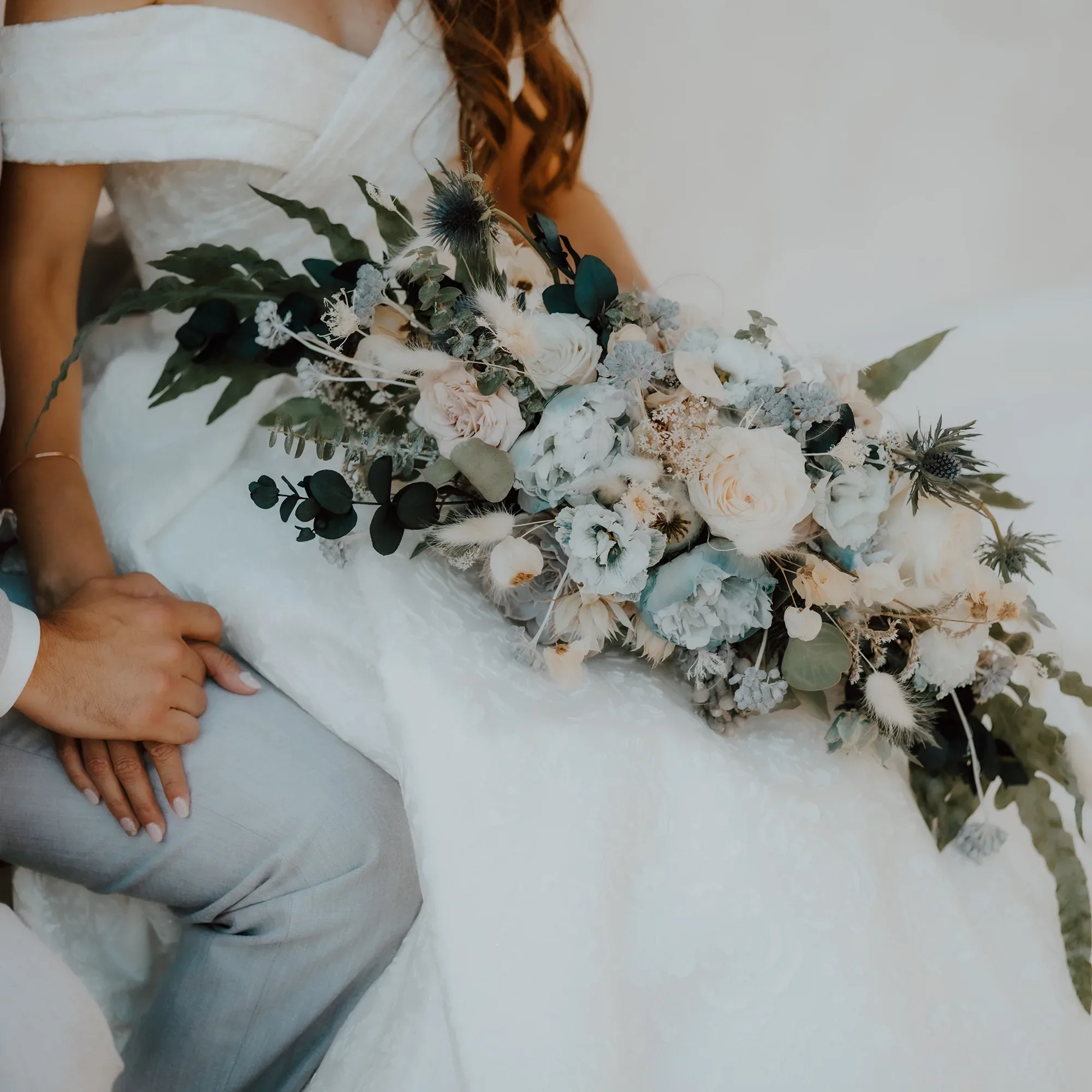 Eine Braut in einem schulterfreien weißen Kleid hält einen opulenten Brautstrauß aus pastellfarbenen Blumen und grünen Blättern, während sie die Hand ihres Partners hält.