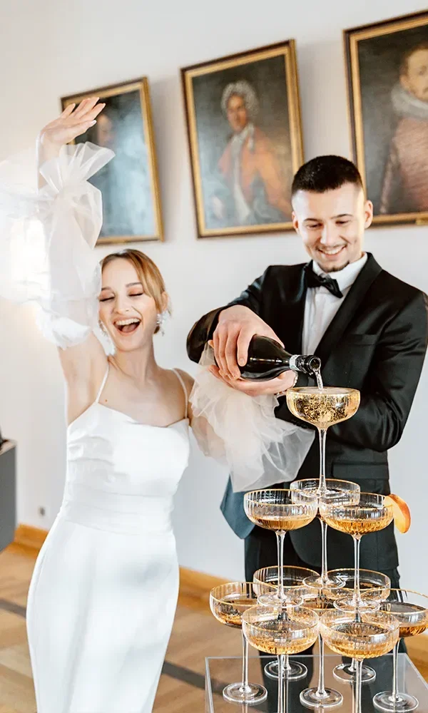 Ein elegantes Paar, die Braut in einem weißen Kleid und der Bräutigam im schwarzen Anzug, gießt fröhlich Champagner in einen pyramidenförmigen Turm aus Gläsern. Die Braut hebt begeistert die Hand, während der Champagner von Glas zu Glas fließt.
