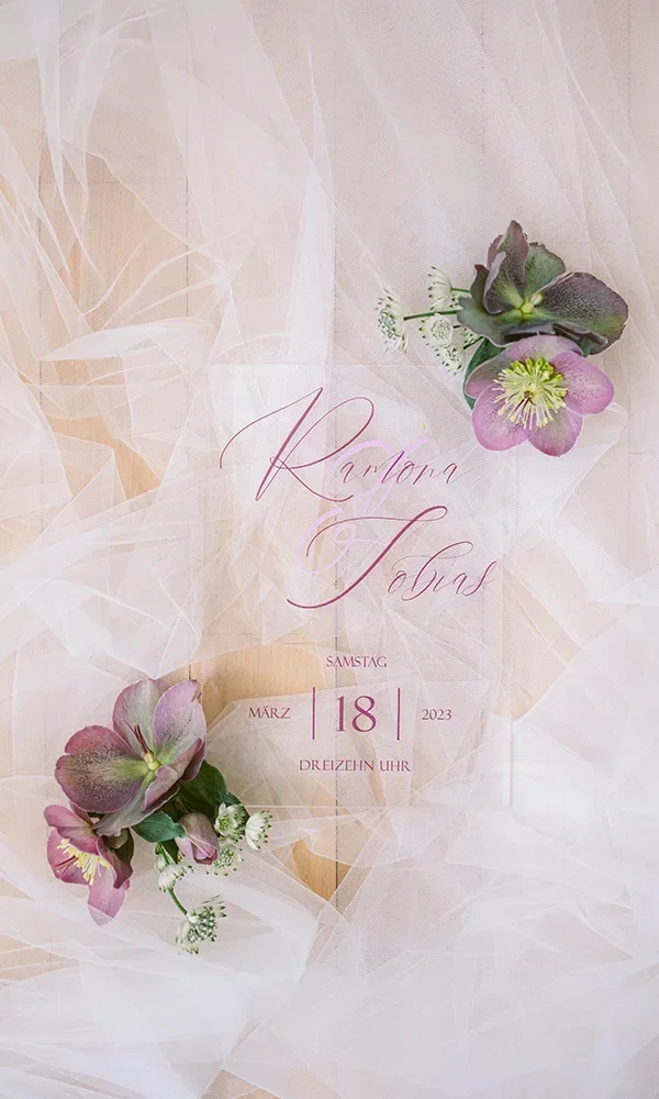 Eine stilvolle Hochzeitseinladung mit den Namen 'Ramona & Tobias' in geschwungener Schrift. Die Einladung ist von lila Blumen und einem zarten, weißen Tüll umgeben und zeigt das Hochzeitsdatum: 18. März 2023.