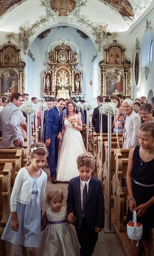 Ein frisch verheiratetes Paar verlässt die Kirche, während Blumenkinder vorausgehen. Die prachtvolle, historische Kirche ist mit Blumenschmuck dekoriert, und die Gäste blicken dem Brautpaar freudig entgegen.