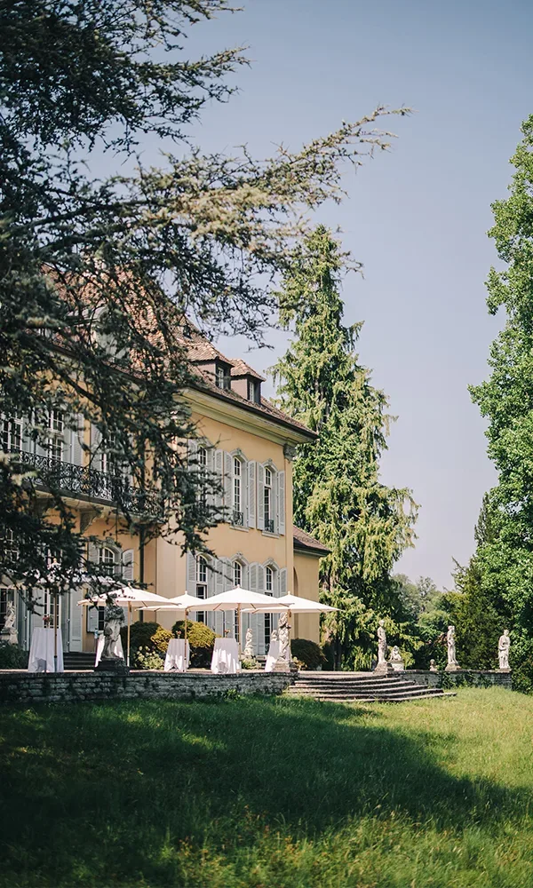 Eine historische Hochzeitslocation im Freien mit gelbem Gebäude, umgeben von Bäumen. Auf der Terrasse stehen festlich dekorierte Tische mit Sonnenschirmen bereit.