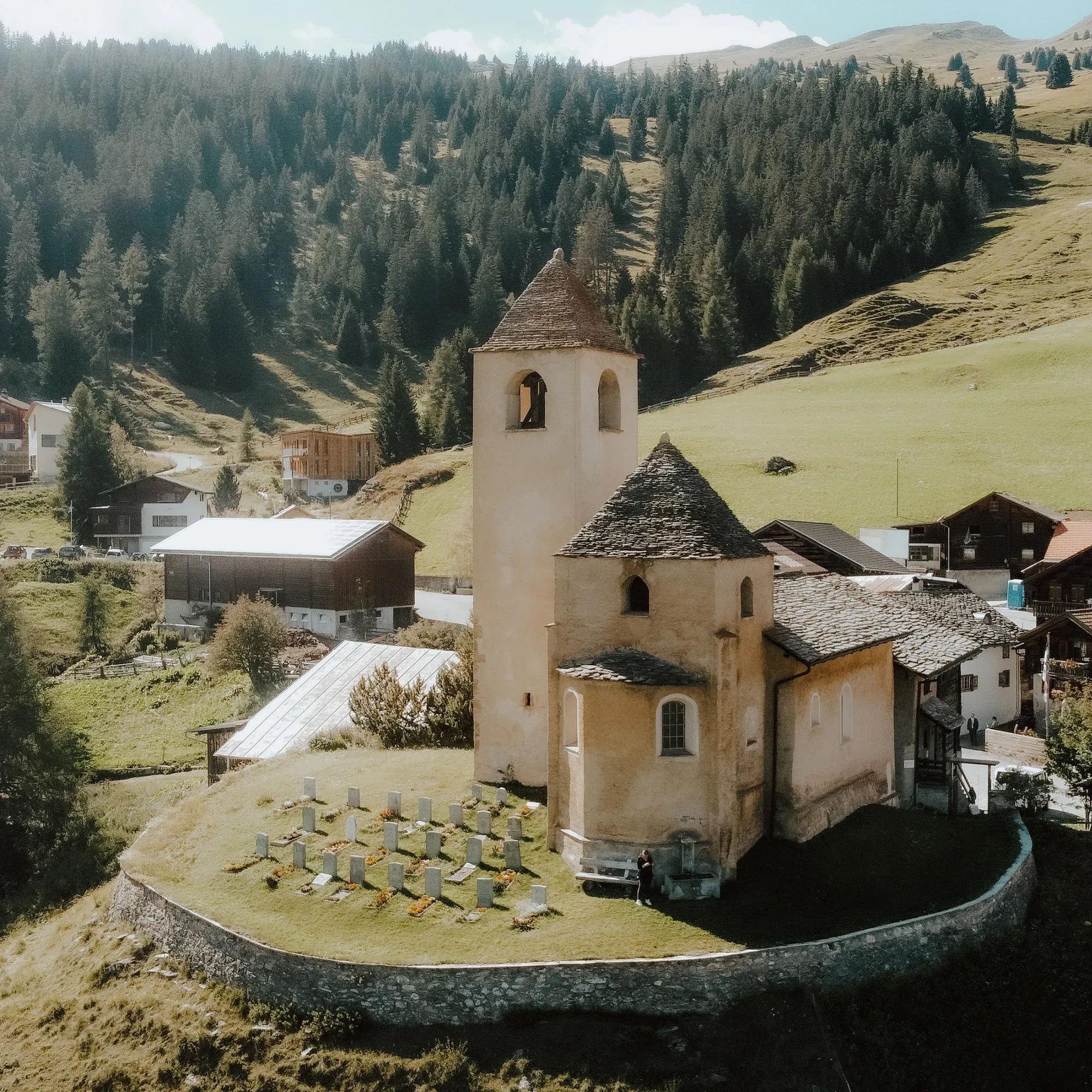 Eine malerische alte Dorfkirche in einer grünen Berglandschaft, umgeben von Wiesen und Holzhäusern, steht auf einem Hügel mit einem kleinen Friedhof.