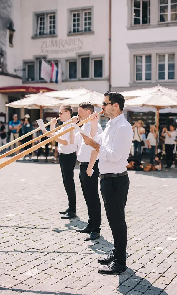 Eine Gruppe von Musikern in weißer Kleidung spielt Alphörner auf einem gepflasterten Platz während einer Hochzeitsfeier. Zuschauer im Hintergrund beobachten die Aufführung.