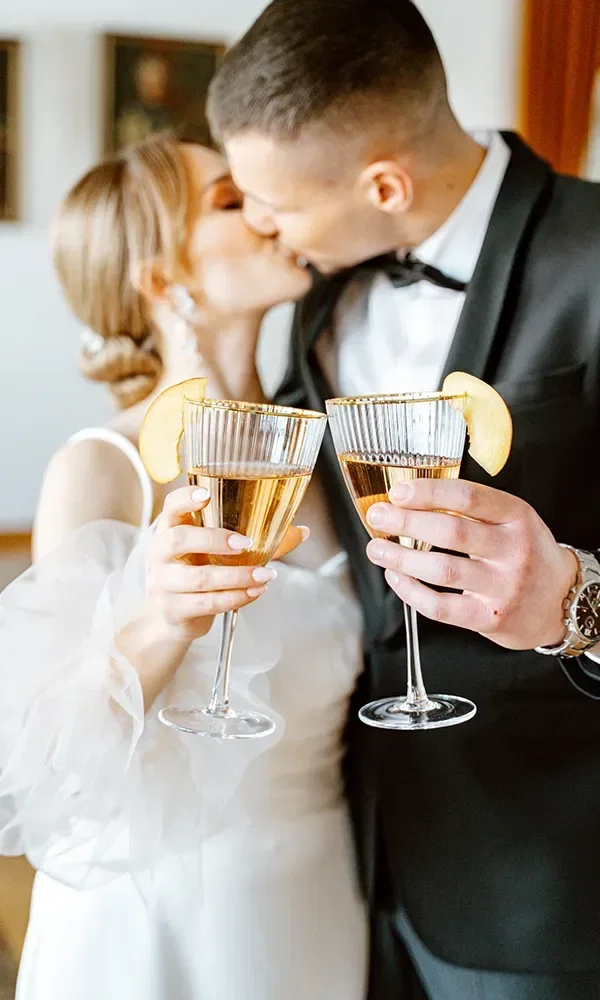 Ein Brautpaar in eleganter Hochzeitskleidung küsst sich zärtlich, während sie zwei mit Champagner gefüllte Gläser halten. Zitronenscheiben verzieren die Gläser, was einen frischen, festlichen Akzent setzt.