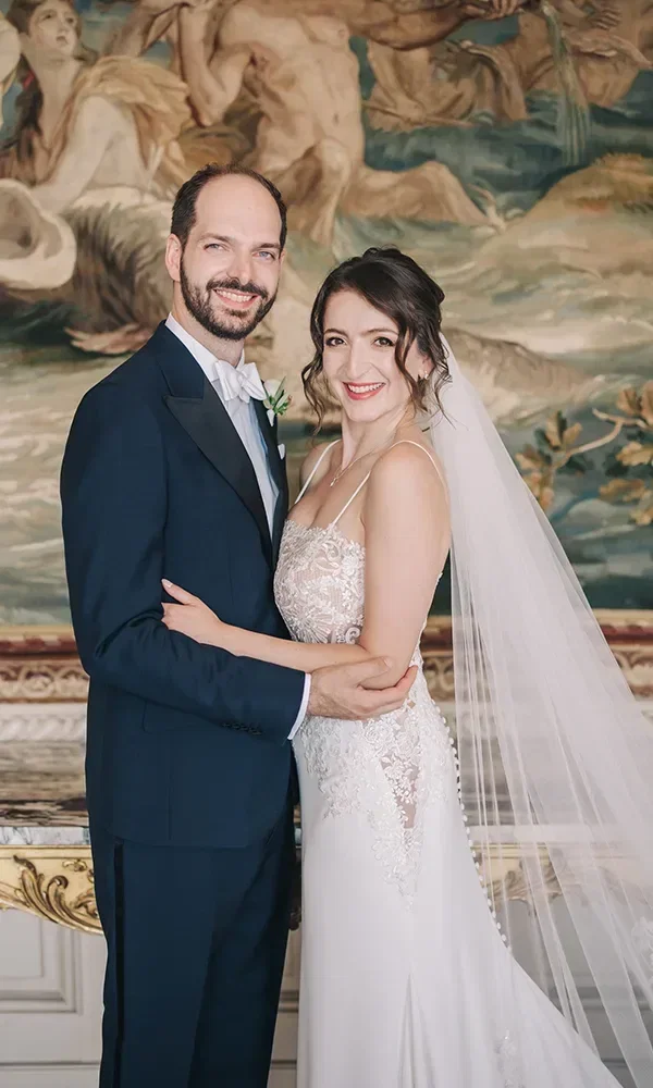 Ein Brautpaar posiert in einer eleganten Umgebung mit einem kunstvollen Gemälde im Hintergrund. Der Bräutigam trägt einen dunklen Anzug, die Braut ein klassisches weißes Kleid mit Schleier.