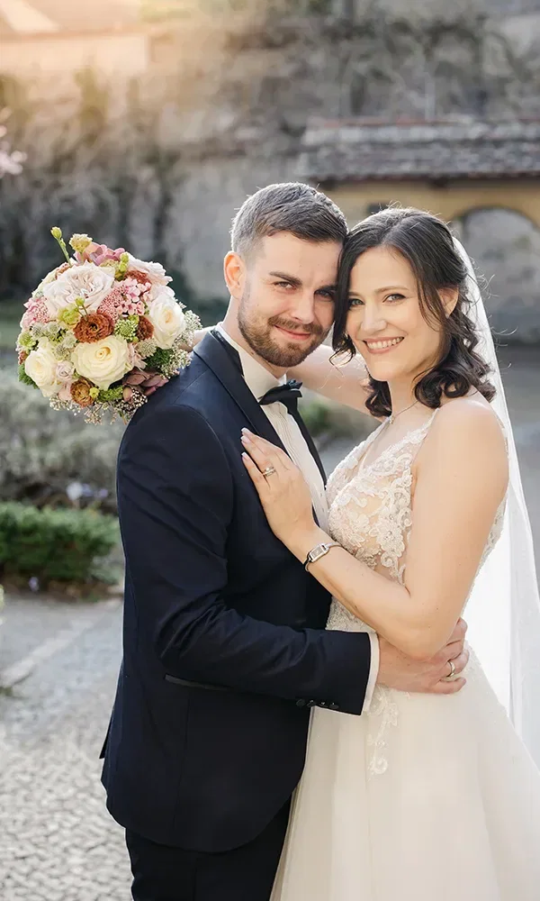 Ein glückliches Brautpaar umarmt sich bei einer Outdoor-Hochzeitsfotosession. Die Braut trägt ein elegantes, ärmelloses Hochzeitskleid und hält einen üppigen Blumenstrauß. Der Bräutigam ist in einem dunklen Anzug mit Fliege gekleidet, und beide lächeln zufrieden in die Kamera.