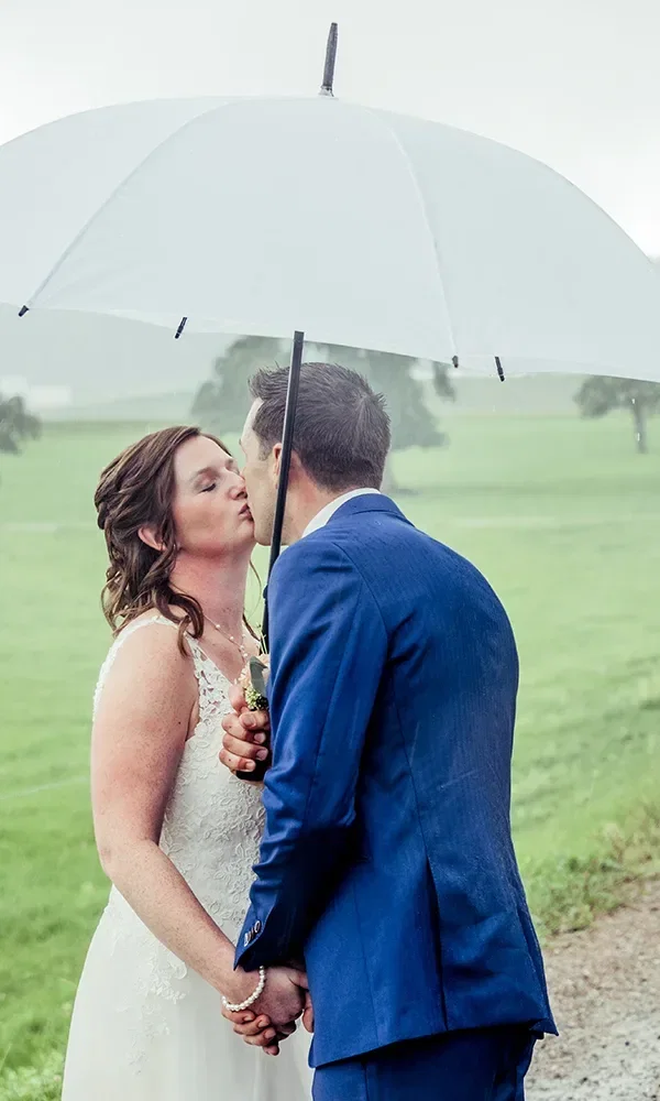 Ein Brautpaar küsst sich unter einem großen weißen Regenschirm bei leichtem Regen auf einer grünen Wiese. Die Braut trägt ein weißes Hochzeitskleid, der Bräutigam einen blauen Anzug.