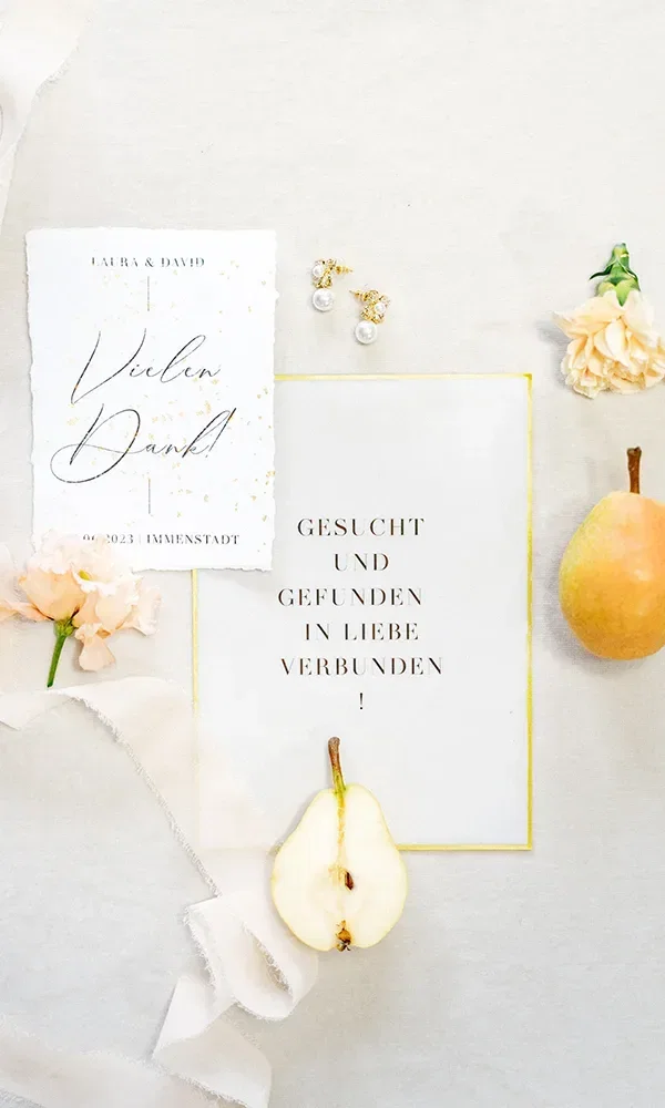 Eine stilvolle Hochzeitseinladung und Dankeskarte, dekoriert mit zarten Blüten und einer aufgeschnittenen Birne, liegen flach auf einer hellen Oberfläche. Kleine goldene Ohrringe ergänzen das Arrangement, das eine elegante und natürliche Stimmung vermittelt.